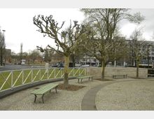 Ein kleiner Park mit blattlosen Bäumen, grünen Sitzbänken und gepflasterten Wegen, eingefasst von einem weißen Metallgeländer. Im Hintergrund sind eine Straße mit Autos sowie Gebäude zu sehen.