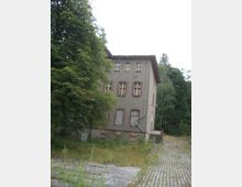 Ansicht eines verlassenen, mehrstöckigen Gebäudes mit grauer Fassade und roten Backsteinkanten um die Fenster. Im Vordergrund befindet sich ein überwucherter gepflasterter Weg, umgeben von Bäumen und wildem Gras.