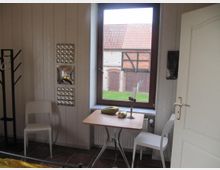 Ein kleiner Raum mit weiß vertäfelten Wänden, einem Fenster mit Blick auf ein Fachwerkgebäude, einem Holztisch mit zwei weißen Stühlen sowie einem Kerzenhalter und einem Buch darauf. Ein weißer Türrahmen und ein Garderobenständer sind ebenfalls sichtbar.