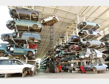 Ein großer Autoverwertungsplatz mit mehreren gestapelten Reihen von Autos ohne Reifen unter einem offenen Hallendach. Ein Gabelstapler hebt ein Auto in die obere Reihe.