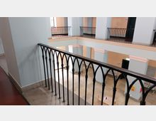 Innenraum eines Gebäudes mit offener Galerie entlang eines Atriums. Zu sehen ist ein Metallgeländer mit bogenförmigen Elementen, helle Wände in Pastellfarben und Säulen mit dekorativen Details.