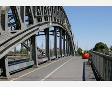 Eisenbrücke mit genieteter Stahlkonstruktion und Straßenbeleuchtung, die über eine Straße mit Fahrrad- und Gehweg führt. Auf der rechten Seite stehen Fahrräder an einem Metallgeländer, im Hintergrund sind Bäume und ein rotes Kunstobjekt zu sehen.