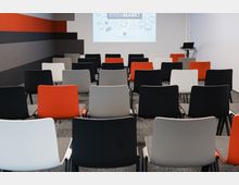 Ein moderner Konferenzraum mit Reihen bunter, minimalistischer Stühle (schwarz, grau, weiß, rot) vor einer Projektionsleinwand, auf der "IDEENMARKT" steht. Rechts steht ein kleiner Tisch mit einem Laptop, die Wände sind schlicht gestaltet.