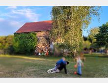Eine alte Scheune mit rotem Ziegeldach und einer teilweise mit Efeu bewachsenen Fassade steht auf einer Wiese. Im Vordergrund spielen Personen auf dem Gras, während Bäume und Sträucher die ländliche Umgebung einrahmen.