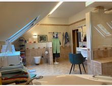 Ein geräumiges Badezimmer mit beige-braunen Fliesen und einer Dachschräge, die ein großes Dachfenster enthält. Zu sehen sind eine freistehende Badewanne im Vordergrund, ein Waschbecken mit Spiegel, ein WC, Handtuchhalter und ein Stuhl; im Hintergrund befinden sich Regale und eine geöffnete Tür.