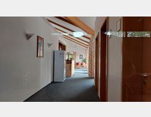 Flur mit Teppichboden, weißen Wänden und schräger Holzdecke mit Sichtbalken, links an der Wand ein Kühlschrank und ein Bild, vorne ein offener Bereich mit Tisch, Stühlen und großen Fenstern, durch bunte Vorhänge eingerahmt.