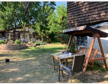 Ein rustikaler Garten mit einem Holztisch, einer Schaukelbank und einem Gartenstuhl im Vordergrund. Im Hintergrund steht ein kleines Holzhaus mit dekorativen Schildern und Pflanzen, umgeben von Bäumen und einem Blumenbeet mit roten Rosen.