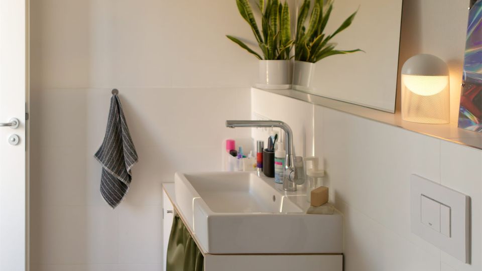 Helles, modernes Badezimmer mit weiß gefliesten Wänden, kleinem Mosaikboden und Waschtisch mit großem Spiegel. Neben dem Waschbecken stehen Pflanzen und Pflegeprodukte; im Vordergrund ist eine Toilette zu sehen, dazu ein Wandregal und ein Handtuchhaken.