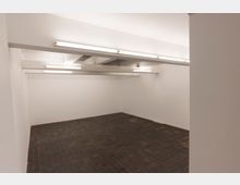 Leerer Raum mit weißen Wänden, dunklem gefliestem Boden und mehreren linearen Deckenleuchten. Die Decke hat sichtbare Träger, die den Raum strukturieren.