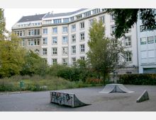 Ein kleiner Skaterpark mit grauen Rampen, die mit Graffiti bemalt sind, umgeben von einer Grünfläche mit Bäumen und Sträuchern. Im Hintergrund steht ein mehrstöckiges Wohn- oder Bürogebäude mit heller Fassade und zahlreichen Fenstern.