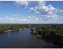 Luftaufnahme eines Flusses, der durch eine dichte Waldlandschaft fließt. Am Ufer befinden sich kleine Anlegestellen mit Booten, umgeben von Bäumen, und im Hintergrund ist eine weite Waldfläche unter einem teilweise bewölkten Himmel zu sehen.