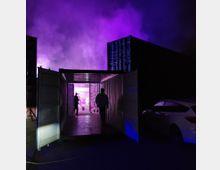 Ein geöffneter Schiffscontainer in einer dunklen Umgebung, beleuchtet von violettem und blauem Licht, mit Personen, die im Inneren stehen. Im Hintergrund sind dichter Rauch oder Nebel und weitere Containerstrukturen sichtbar, rechts daneben ein parkendes weißes Auto.