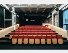 Kleiner Veranstaltungssaal mit mehreren Reihen roter Klappsitze und einem dunklen Bühnenbereich im Hintergrund. Der Raum hat industrielle Wände, sichtbare Kabel und eine minimalistische Deckenbeleuchtung.