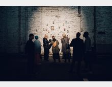 Eine Gruppe von Personen steht in einem dämmrig beleuchteten Raum vor einer weißen Backsteinwand, an der kleine Porträtbilder angebracht sind. Die Szenerie wirkt wie eine Ausstellung in einem industriellen oder künstlerischen Ambiente.