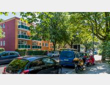 Mehrfamilienhaus mit farbigen Fassaden (orange und rot) und Balkonen mit grünen Brüstungen entlang einer von Bäumen gesäumten Straße. Geparkte Autos und Motorroller stehen am Straßenrand, während im Hintergrund ein Lieferwagen vor einem Straßenschild ("Elisenstraße") sichtbar ist.