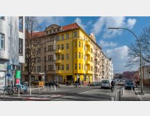 Straßenszene in einer Stadt mit mehrstöckigen Wohnhäusern, darunter ein auffälliges, gelbes Gebäude mit Balkonen. Im Vordergrund fahren Autos, Fahrräder und Fußgänger auf einer Straße mit Verkehrsschildern und Fahrradständern.