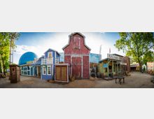 Panorama einer Westernstadt-Kulisse mit rustikalen Gebäuden, darunter eine rote Scheune, ein blaues Holzhaus, ein Saloon mit Holzveranda und ein Sheriff-Büro. Im Hintergrund steht eine Windmühle und eine blaue Kuppel, umgeben von Bäumen und sandigem Boden.