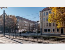Straßenszene in einer Stadt mit mehrstöckigen Wohngebäuden aus heller Fassade und rotem Ziegeldach. Im Vordergrund verlaufen Straßenbahnschienen, Fußgängerwege und Geländer, im Hintergrund stehen Bäume mit herbstlichem Laub.