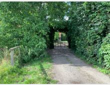 Ein schmaler, unbefestigter Weg führt durch dichte grüne Vegetation zu einem Holztor, das von einem Bogen aus Pflanzen umgeben ist. Im Hintergrund ist eine offene Fläche mit weiteren Bäumen und einem Gebäude sichtbar.