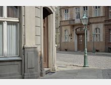 Straßenszene mit historischen Gebäuden aus sandfarbenem Mauerwerk, hohen Fenstern und dekorativen Türen. Pflastersteine bedecken den Gehweg und die Straße; eine grüne, altmodische Straßenlaterne steht im Vordergrund.