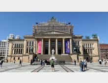 Das Bild zeigt das Konzerthaus Berlin auf dem Gendarmenmarkt, eine neoklassizistische Fassade mit Säulen und einer Statue im Eingangsbereich. Baugerüste bedecken Teile des Dachs, während bunte Banner an den Säulen hängen; der Vorplatz ist von Besuchern bevölkert.