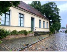 Straßenszene mit einem historischen Gebäude aus hellgelbem Putz und grünen Fensterrahmen. Das Haus steht an einer gepflasterten Straße, umgeben von Bäumen und kleinen Vorgärten.