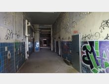 Ein verlassener Gang mit abblätterndem Putz an den Wänden, blauen Fliesen im unteren Bereich und mehreren Graffiti. Die Decke zeigt freiliegende Leitungen, und im Hintergrund führen Türöffnungen in dunklere Bereiche.