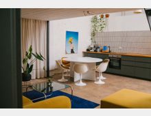 Offene Wohnküche mit weißer Backsteinwand, langer Küchenzeile mit dunklen Schränken und Holzarbeitsplatte sowie hell gefliestem Spritzschutz. In der Mitte steht ein ovaler weißer Esstisch mit vier Stühlen; daneben hängen Pflanzen von der Decke, ein Vorhang trennt den Raum und im Vordergrund liegt ein blauer Teppich mit Glastisch.