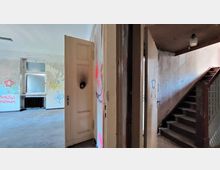 Ein verlassener Innenraum eines Gebäudes mit abgenutzten Wänden, Graffiti und einer offenen Holztür. Rechts führt eine schmale Holztreppe nach oben, links ist ein Raum mit einer Heizkörper-Nische und einem Fenster zu sehen.