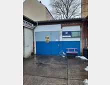 Außenansicht einer Kfz-Werkstatt mit blauer Metalltür, teilweise weiß-blauer Wand und einem Firmenschild. Ein pinkfarbener Holzbänkchen steht rechts neben der Tür, dahinter ein großer Schornstein aus Backstein und ein Baum im Hintergrund.