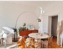 Helles Wohnzimmer mit einem runden Esstisch aus weißem Marmor, umgeben von Holzstühlen. Im Hintergrund stehen eine Holzkommode mit Zimmerpflanzen und Bildern sowie eine weiße Tür mit Stuckdetails an der Decke, ergänzt durch eine elegante Bogenlampe und Dekorationen.