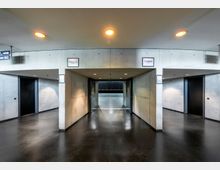 Modernes Innenraumdesign eines Gebäudes mit Betonwänden und dunklem, poliertem Boden. Der Raum zeigt einen offenen Übergang mit mehreren schwarzen Türen und darüber angebrachten Schildern.