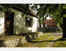 Ein kleines Gebäude mit weiß gestrichenen Ziegelwänden und dunklen Gittern vor einer Holztür und den Fenstern steht im Vordergrund, umgeben von Bäumen. Im Hintergrund ist ein Haus mit roten Ziegeldächern zu sehen, umgeben von einer niedrigen Steinmauer.