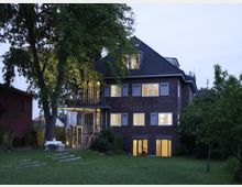 Backsteinhaus mit mehreren beleuchteten Fenstern in einem großen Garten bei Dämmerung. Vor dem Haus stehen ein großer Baum und ein gedeckter Tisch mit Stühlen, umgeben von Rasenflächen und weiteren Bäumen.