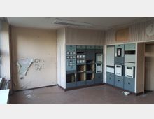 Verlassener Raum mit technischen Schaltkästen an der Wand, beschädigtem Putz und abblätternder Wandfarbe. Die Einrichtung wirkt altmodisch, und der Raum scheint in Verfall geraten zu sein.