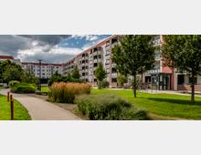 Mehrstöckiges Wohngebäude mit roten Dachelementen und Balkonen, umgeben von einer gepflegten Grünanlage mit Rasen, Bäumen, Ziergräsern und einem gepflasterten Weg, der durch die Anlage führt. Im Vordergrund stehen Fahrräder und ein überdachter Eingangsbereich.