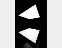 Zwei große, weiße, dreieckige Lichtflächen leuchten in einem dunklen Raum und sind übereinander positioniert, was einen minimalistischen und geometrischen Effekt erzeugt. Die Umgebung ist dunkel und hebt die hellen Flächen hervor.