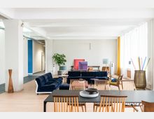 Heller, moderner Wohnraum mit offenen Bereichen, bestehend aus einem Essbereich mit schwarzem Tisch und hölzernen Stühlen sowie einer Sitzecke mit blauem Sofa und dunkelblauen Sesseln. Große Fenster rechts lassen viel Tageslicht herein; an der Wand stehen Kunstwerke und dekorative Objekte, darunter ein Blumentopf und eine Stehlampe.