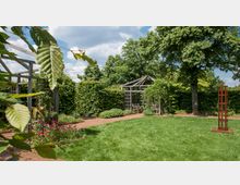 Ein gepflegter Garten mit einer Rasenfläche, Blumenbeeten und einem Weg, der zu einer überdachten Pergola aus Holz führt. Im Hintergrund stehen dichte grüne Hecken und Bäume, während rechts eine moderne, rustikale Skulptur aus Metall zu sehen ist.