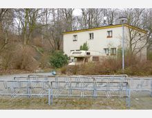 Freistehendes zweistöckiges Wohnhaus mit heller Fassade und gelben Dachkanten, umgeben von winterlichen, blattlosen Bäumen und Büschen. Im Vordergrund befindet sich ein Fahrradständer auf einem gepflasterten Platz, daneben ein geparktes Wohnmobil.