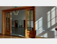 Ein Innenraum mit einer großen doppelflügeligen Holztür mit Glaseinsätzen, die in einen anderen Raum mit grauem Bodenbelag und einem modernen Kronleuchter führt. Sonnenlicht fällt durch ein Fenster auf die weiße Wand und den Parkettboden im Vordergrund.