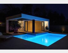 Modernes, flaches Pavillongebäude mit breitem Vordach und großen bodentiefen Glasfronten, nachts warm beleuchtet. Davor liegt ein rechteckiger, blau beleuchteter Swimmingpool mit umlaufender Terrasse; im Hintergrund sind Bäume und ein Sichtschutzzaun zu sehen.