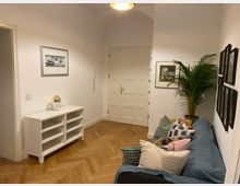 Ein gemütlich eingerichtetes Wohnzimmer mit hellen weißen Wänden und einem Holzboden im Fischgrätenmuster. Zu sehen sind ein blaues Sofa mit bunten Kissen, eine weiße Kommode mit Dekoration, mehrere Bilderrahmen an den Wänden und eine große grüne Zimmerpflanze in einer Ecke.