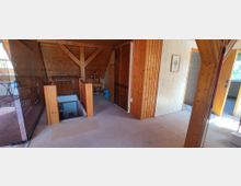 Dachgeschoss eines Hauses mit sichtbaren Holzbalken und Holzverkleidung an Wänden und Decke. Zu sehen sind ein Treppenabgang mit Geländer, ein Wandspiegel, Türen zu angrenzenden Räumen sowie ein schmales Fenster, das Tageslicht hereinlässt.