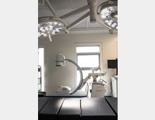 Moderner Operationssaal mit zwei großen OP-Leuchten an der Decke, einem C-Bogen-Röntgengerät und weiteren medizinischen Geräten entlang der Wände. Im Vordergrund ist eine schwarze Patientenliege zu sehen.