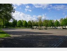 Weitläufiger, gepflasterter Platz mit vereinzelten Baumreihen und angrenzenden Grünflächen; im Hintergrund sind Wohngebäude und parkende Autos zu sehen, unter einem Himmel mit leichter Bewölkung.