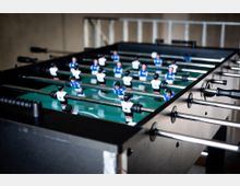 Ein Tischkicker in einem Innenraum, mit grünem Spielfeld, Metallstangen und Spielfiguren in blauen und weißen Trikots. Der Tisch hat ein dunkles Gehäuse und ist aus schräger Perspektive aufgenommen, sodass die Reihen der Figuren und Stangen in die Tiefe verlaufen.