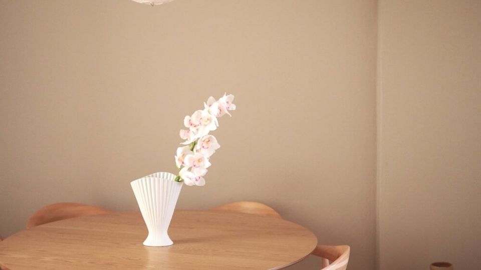 Minimal dining nook with a round wooden table and four light wood chairs on a dark wood plank floor, set against plain beige walls. A white paper pendant lamp hangs overhead, and a vase with pale flowers sits centered on the table.