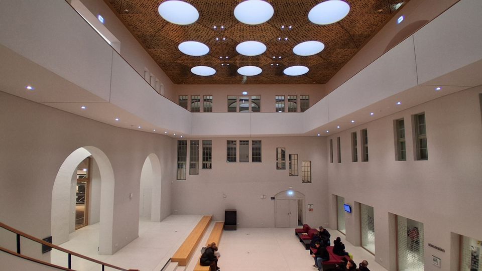 Großes, modernes Atrium mit hellen Wänden, umlaufenden Galerien und vielen rechteckigen Fenstern auf mehreren Ebenen. Die Decke ist mit runden Oberlichtern und einer perforierten Holzverkleidung gestaltet; im Erdgeschoss stehen Bänke und Sitznischen neben hohen Rundbögen und Glasfronten.