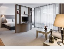 Zimmer in modernem, minimalistischem Design mit einer halbhohen Trennwand, auf der ein Flachbildfernseher steht. Im Hintergrund befindet sich ein Bett mit grauem Kopfteil, im Vordergrund ein Couchtisch mit Glasplatte und dekorativen Elementen, sowie bodentiefe, halbtransparente Vorhänge an großen Fenstern.